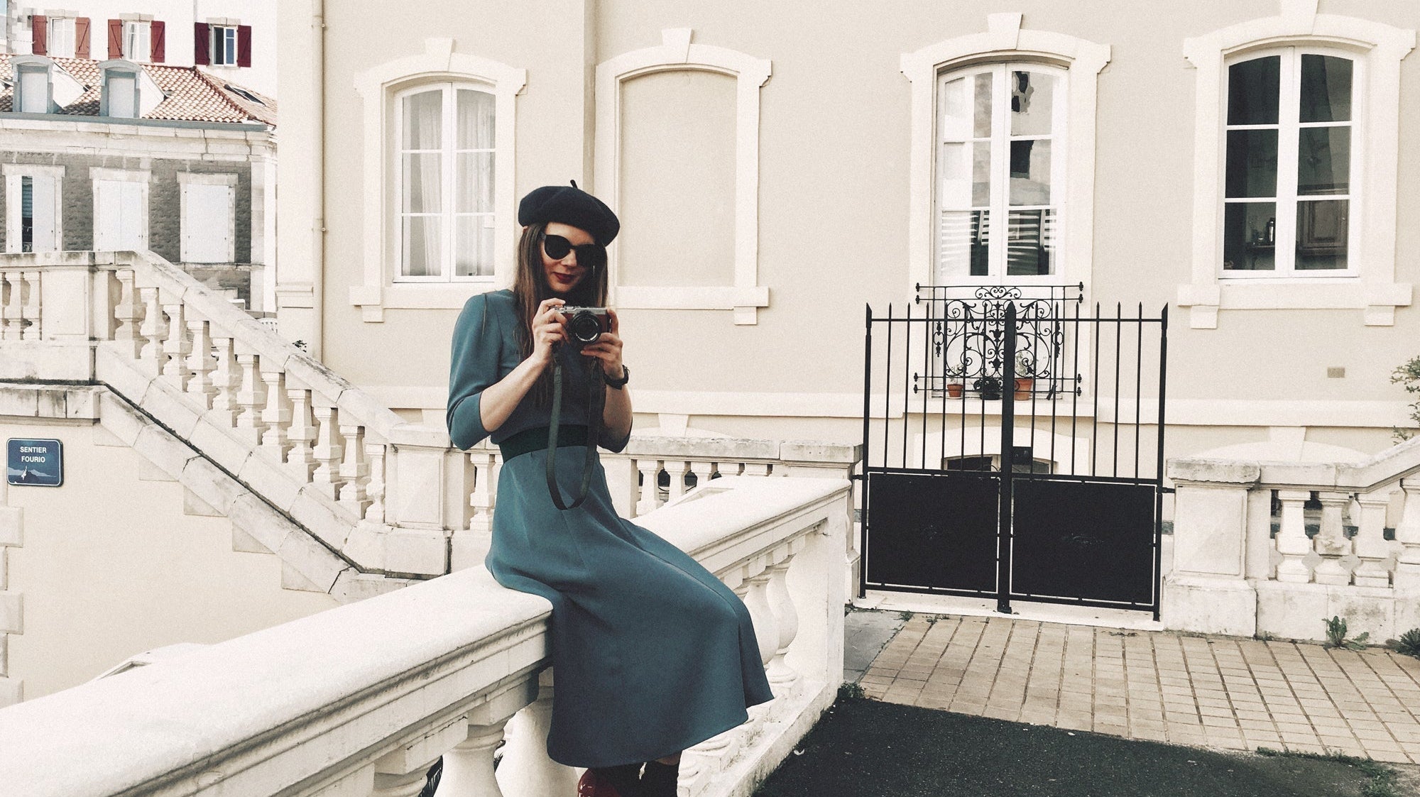 Person sitting on a street wall holding a camera wearing a blue dress and beret in Biarritz, France.