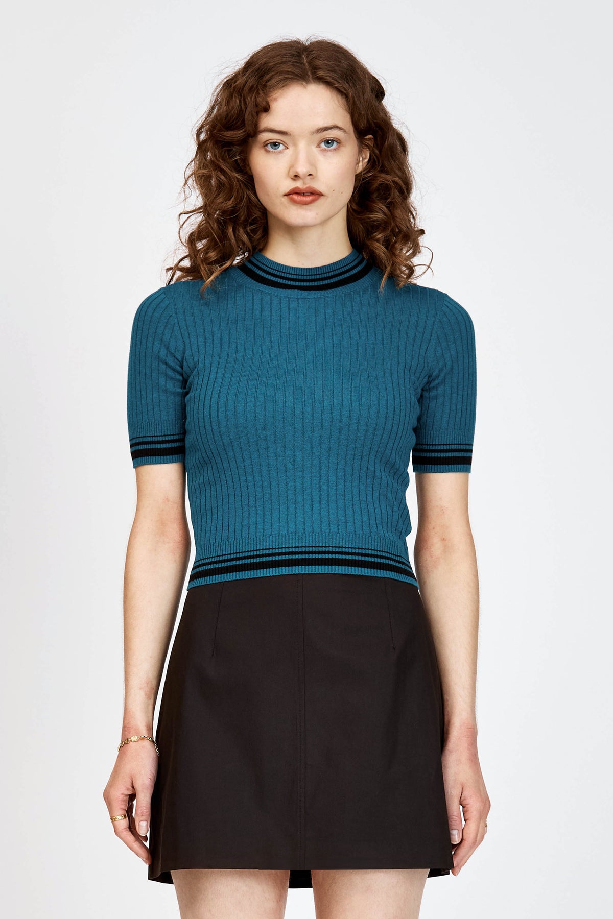 A woman modeling a teal short-sleeve ribbed knit mock turtle neck top with black stripes at the collar, sleeve cuffs, and hem, paired with a black skirt, against a light gray studio background.