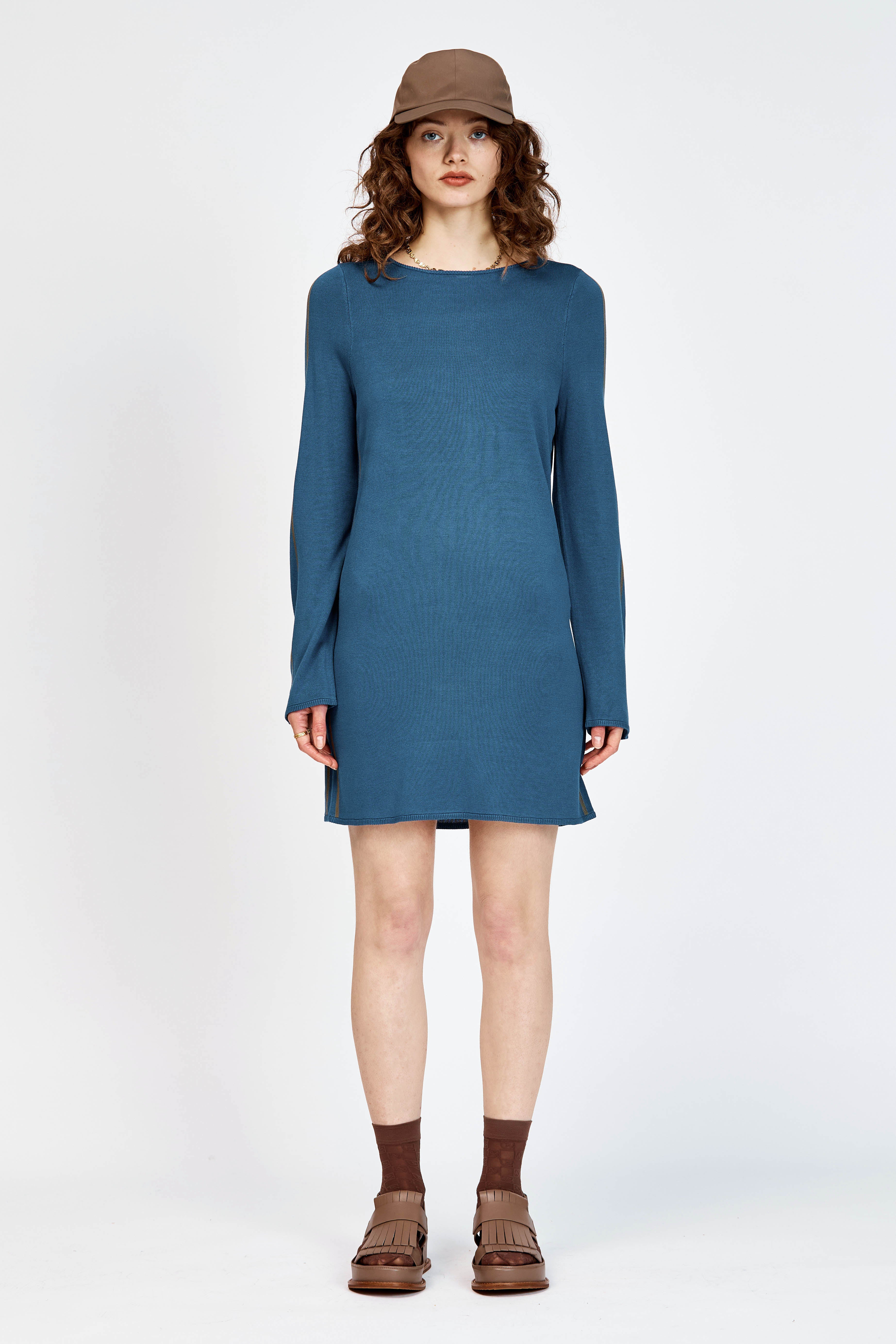A model wearing a slate blue long-sleeve knit dress with brown side stripes, styled with a brown baseball cap, brown socks, and brown leather sandals against a plain white studio background.