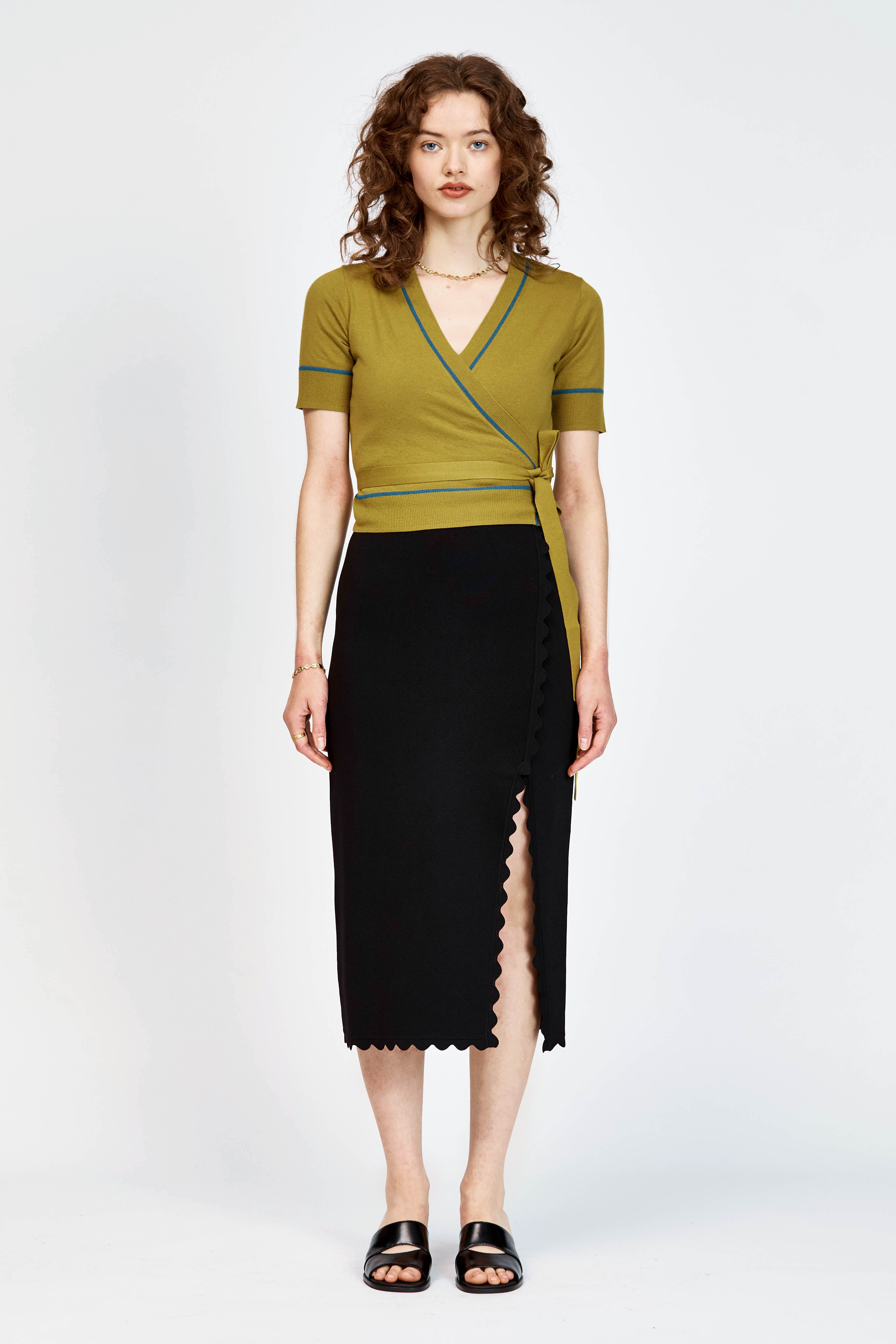 A woman modeling a short-sleeve wrap-style knit top in green with thin teal-blue piping details along the V-neck, sleeve edges, and waist tie, paired with a black skirt against a light gray studio background.