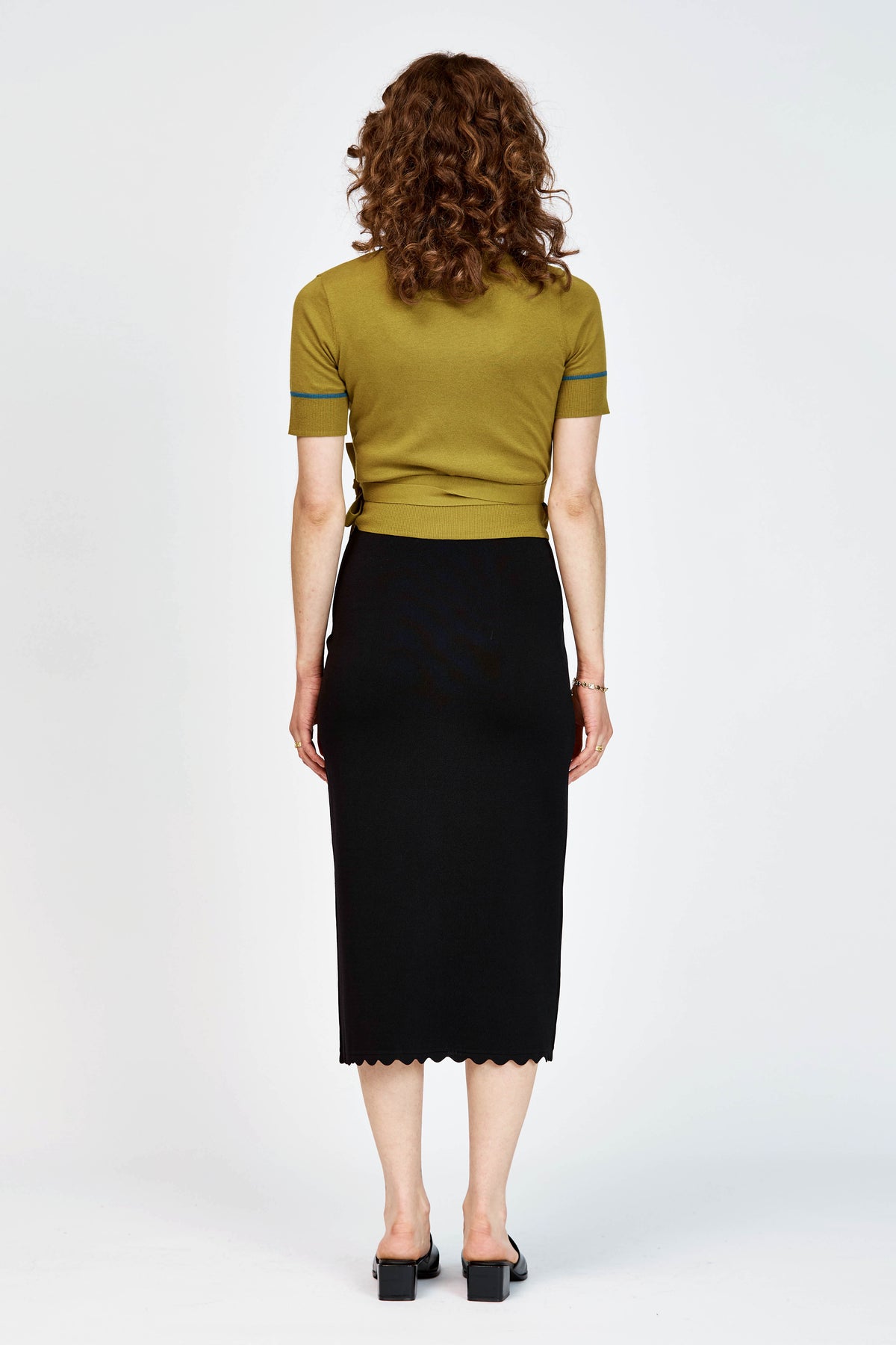 A woman modeling a short-sleeve wrap-style knit top in green with thin teal-blue piping details along the V-neck, sleeve edges, and waist tie, paired with a black skirt against a light gray studio background.