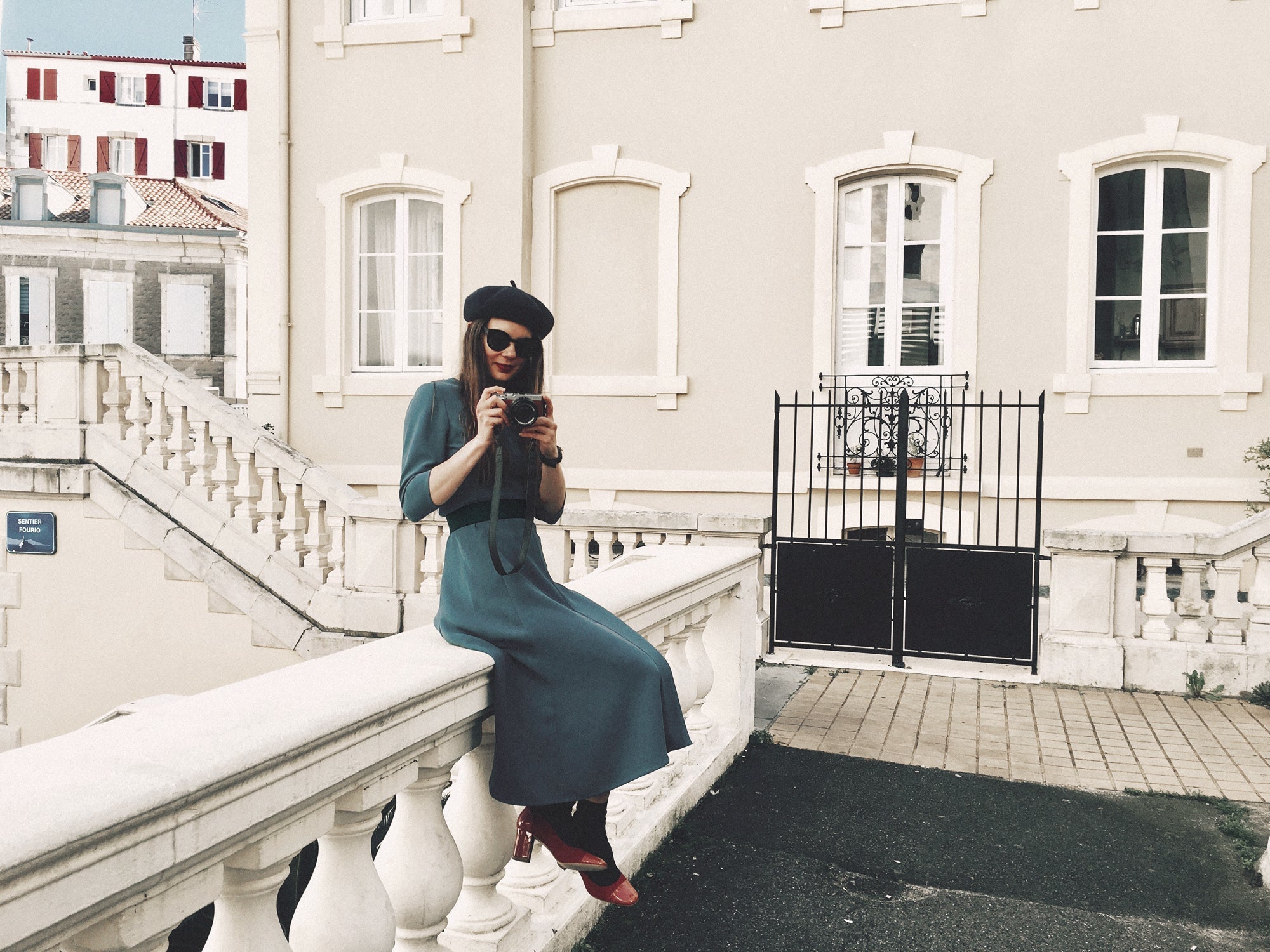 Person sitting on a street wall holding a camera wearing a blue dress and beret in Biarritz, France.