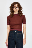 A woman with curly brown hair and glasses wearing a short-sleeved ribbed knit top in burgundy and dark blue jeans, standing against a light gray studio background.