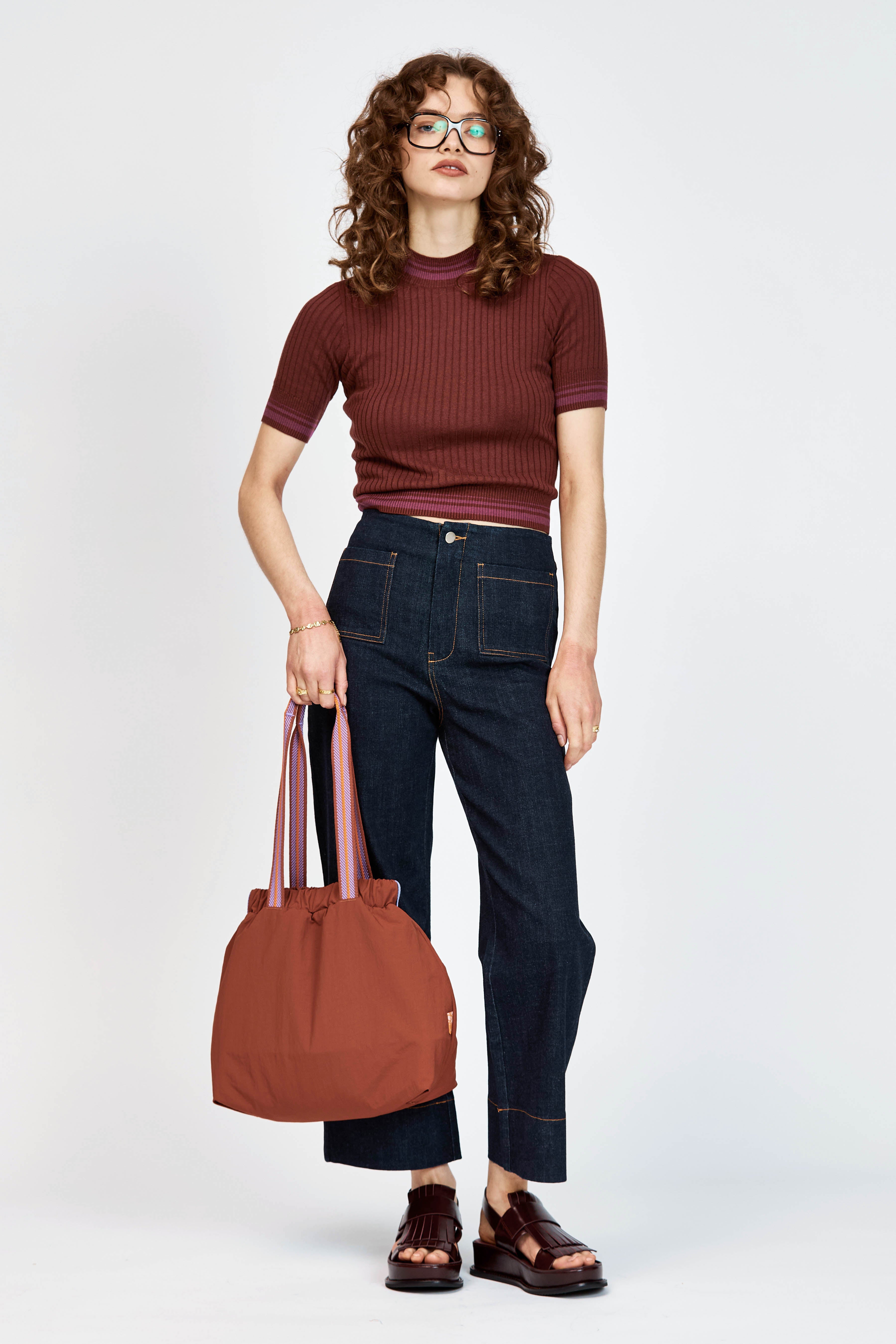 A woman with curly brown hair and glasses wearing a short-sleeved ribbed knit top in burgundy and dark blue jeans, standing against a light gray studio background.