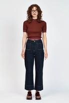 A woman with curly brown hair and glasses wearing a short-sleeved ribbed knit top in burgundy and dark blue jeans, standing against a light gray studio background.