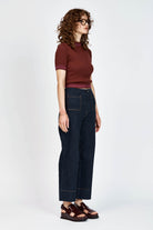 A woman with curly brown hair and glasses wearing a short-sleeved ribbed knit top in burgundy and dark blue jeans, standing against a light gray studio background.