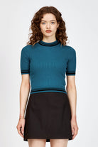 A woman modeling a teal short-sleeve ribbed knit mock turtle neck top with black stripes at the collar, sleeve cuffs, and hem, paired with a black skirt, against a light gray studio background.