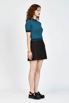 A woman modeling a teal short-sleeve ribbed knit mock turtle neck top with black stripes at the collar, sleeve cuffs, and hem, paired with a black skirt, against a light gray studio background.