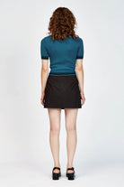 A woman modeling a teal short-sleeve ribbed knit mock turtle neck top with black stripes at the collar, sleeve cuffs, and hem, paired with a black skirt, against a light gray studio background.