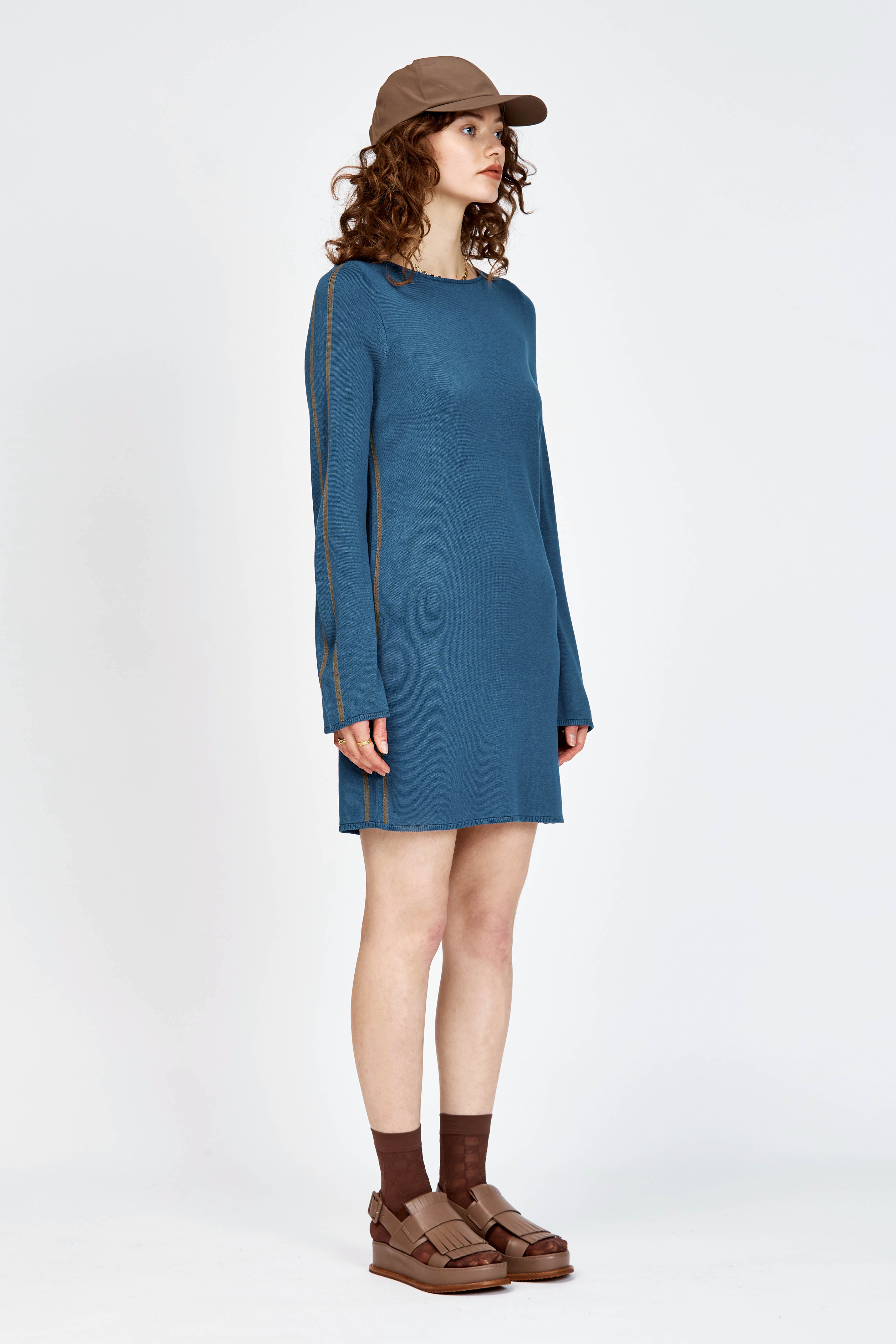 A model wearing a slate blue long-sleeve knit dress with brown side stripes, styled with a brown baseball cap, brown socks, and brown leather sandals against a plain white studio background.