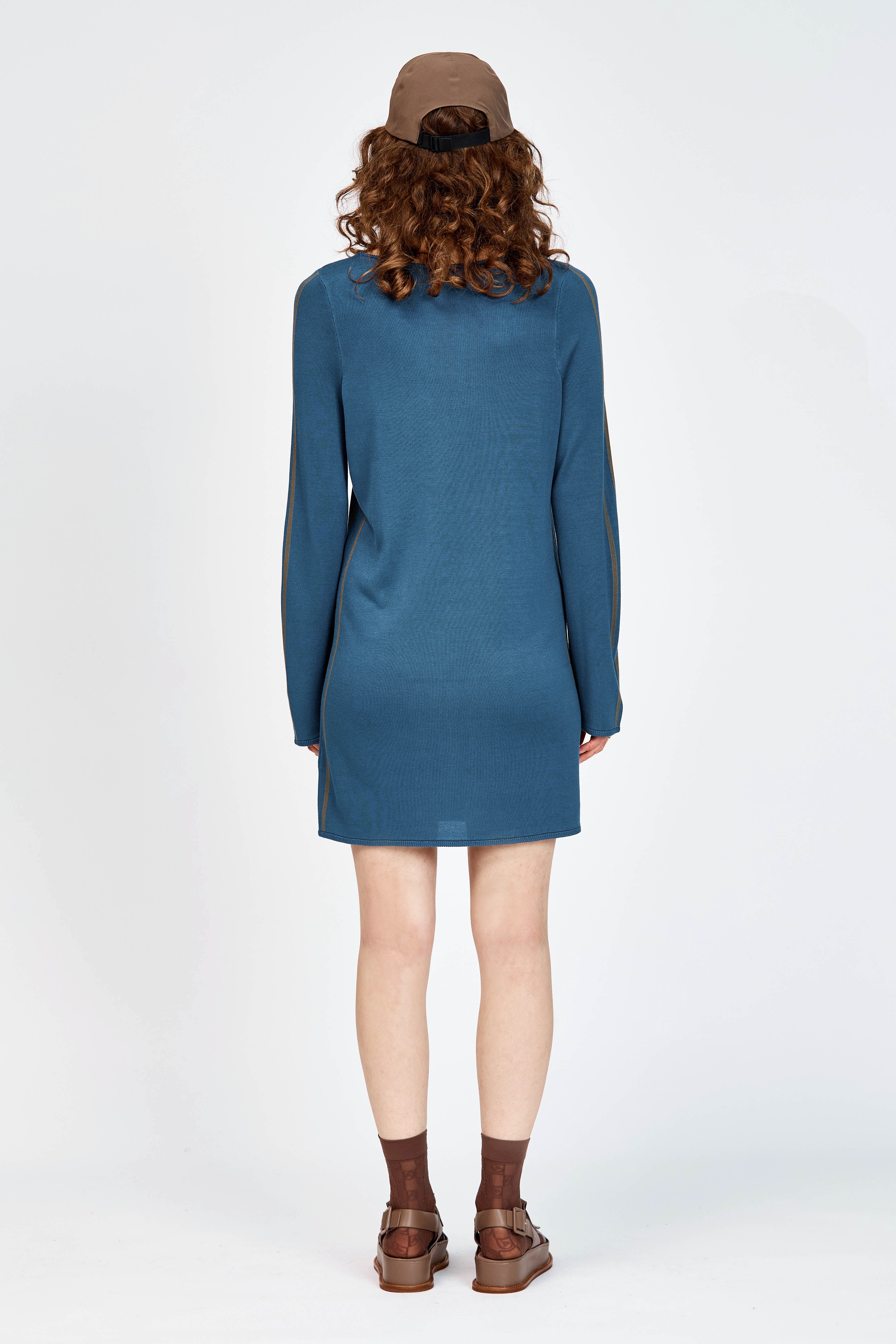 A model wearing a slate blue long-sleeve knit dress with brown side stripes, styled with a brown baseball cap, brown socks, and brown leather sandals against a plain white studio background.