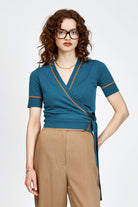 A woman modeling a teal wrap-style short-sleeve top with thin orange trim and tan cropped trousers, standing against a light gray studio background.