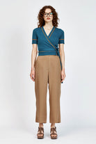 A woman modeling a teal wrap-style short-sleeve top with thin orange trim and tan cropped trousers, standing against a light gray studio background.