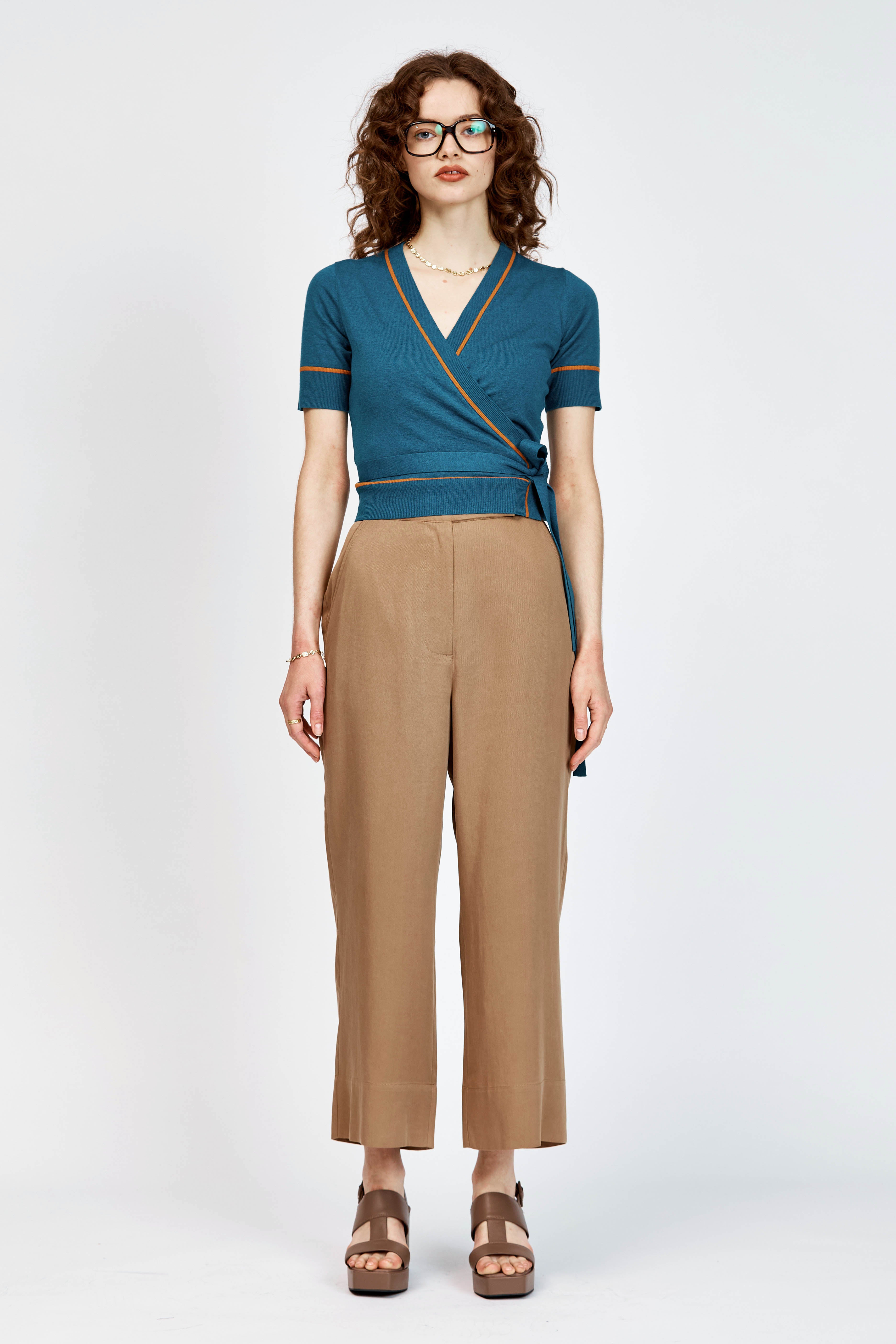 A woman modeling a teal wrap-style short-sleeve top with thin orange trim and tan cropped trousers, standing against a light gray studio background.