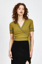 A woman modeling a short-sleeve wrap-style knit top in green with thin teal-blue piping details along the V-neck, sleeve edges, and waist tie, paired with a black skirt against a light gray studio background.