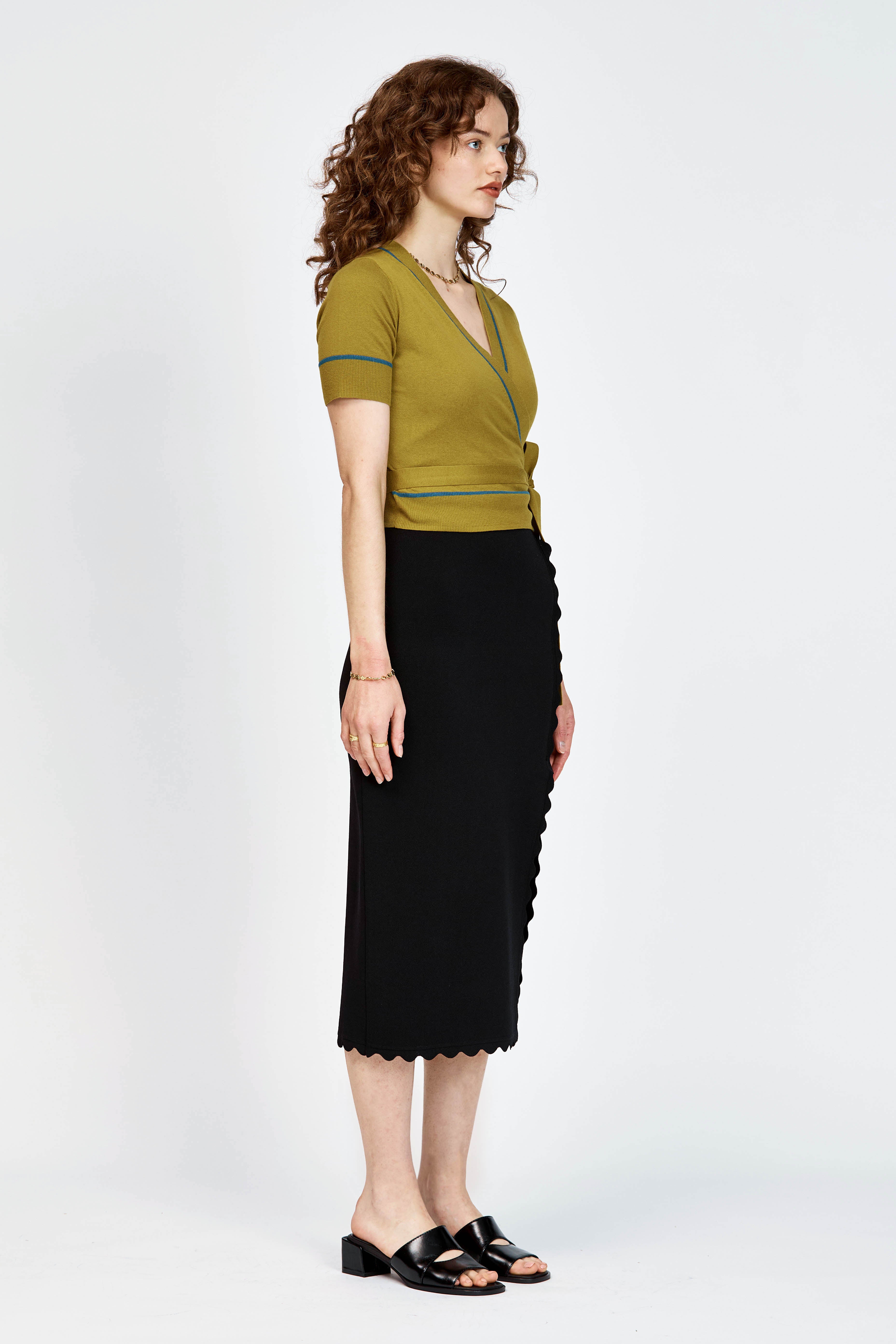 A woman modeling a short-sleeve wrap-style knit top in green with thin teal-blue piping details along the V-neck, sleeve edges, and waist tie, paired with a black skirt against a light gray studio background.