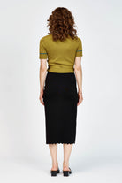 A woman modeling a short-sleeve wrap-style knit top in green with thin teal-blue piping details along the V-neck, sleeve edges, and waist tie, paired with a black skirt against a light gray studio background.
