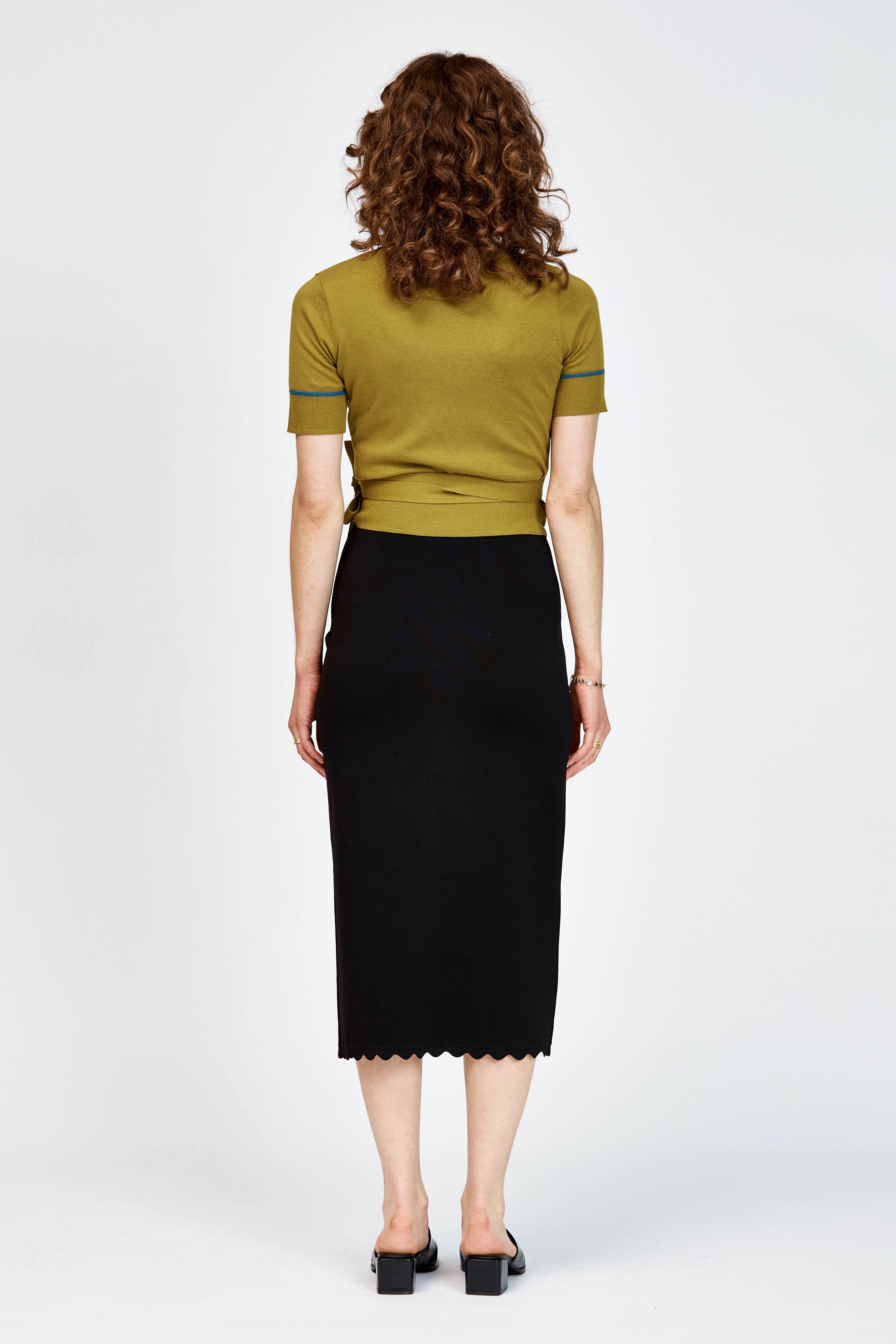 A woman modeling a short-sleeve wrap-style knit top in green with thin teal-blue piping details along the V-neck, sleeve edges, and waist tie, paired with a black skirt against a light gray studio background.